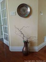 Full view of metal pitcher with faux branch arrangement and metal plate mounted on wall.