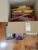 Top view of a cardboard box filled with loose tapered candles in multiple colors including yellow, red, pink, and green. Also visible are the tops of two Colonial Candle boxed sets.