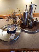 Photo showing stainless steel tea pot, coffee pot, creamer, sugar bowl on a large round serving tray, with another large tray and small dish in foreground, and decorative vase with dried foliage.