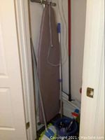 Ironing board covered with beige fabric standing behind several cleaning tools including mops and handles in a closet