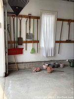 Wide view showing snow shovel hanging on wall along with garden rakes, shovel, broom, fan rake, and planters on floor.