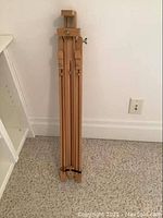Folded wooden easel standing upright on carpeted floor near a white shelf and wall outlet.