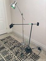 Two metal floor lamps, one silver with vertical pole and one black with horizontal arm, displayed on floor next to wall and carpet.