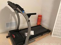Side view of treadmill showing black and gray frame with orange exercise mat rolled up near it.