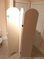 Three white wooden panels of the room divider shown upright and connected by brass hinges, with rounded tops and cutouts at bottoms.