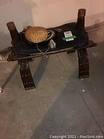 Front view of wooden saddle bench with black leather cushion, woven bag and wicker bottle on seat