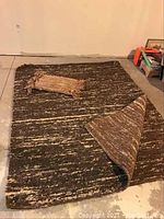 Full view of large brown and gray variegated wool rug with corner flipped, small fringed piece atop.