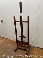 Full view of the vintage wooden artist easel standing on wooden floor against a white wall, showing its general shape and condition.