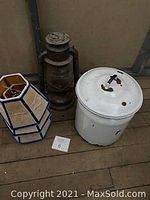 Photo showing vintage enamelled slop jar with lid, antique oil lantern, and three lampshades grouped together on floor.