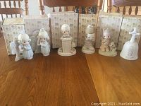 Photo of 6 Precious Moments figurines with six original boxes behind them, showing various children-themed designs and a bell figurine, all with pastel colors and soft finishes.