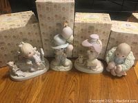 Group of four Precious Moments figurines with matching boxes behind on wooden surface. They depict children in various poses, some with accessories and animals.