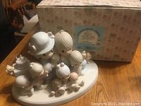 Photo of a multi-piece Precious Moments group figurine on an oval white base with children and small animals, white and pastel colors, alongside original box with Precious Moments branding and graphics.