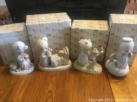 Four Precious Moments figurines in front of their matching boxes on a wooden table. Figurines include children and animals in pastel matte colors.