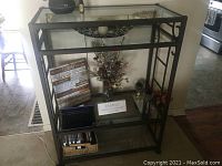 Front view of brown metal shelving unit with three glass shelves, showing approximate dimensions and decorative scrollwork on sides.