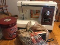 Kenmore sewing machine on wooden table alongside plastic bags of assorted sewing items and large red tin container.