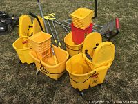 Four yellow Rubbermaid mop buckets equipped with mop wringers and two small yellow buckets, five gray mop handles with yellow grips, and a metal utility cart on grass.