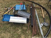 Full overview of lot showing multiple blue and black plastic snow shovels, various metal and wooden handled construction shovels, rusted metal pipes, black flexible hose, and hand tools arranged on grass.
