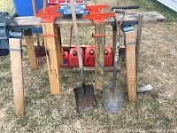 View of two red metal sawhorse brackets attached to wooden legs along with four red gas cans lined up underneath, two shovels standing upright leaning against the sawhorses on grass