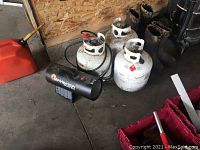 Photo showing black Mr. Heater forced air heater with hose, three white propane tanks, and red gas can on floor.