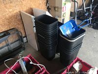 Two stacks of black plastic waste paper baskets next to two white metal Frost garbage containers with angled tops.