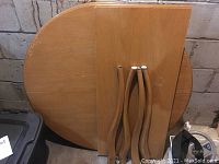 Round birch kitchen table top with extension leaf and 4 detached wooden legs in front, placed on a concrete floor against a block wall.