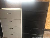 Photo of two metal 3-drawer file cabinets standing side by side against a wooden wall. One cabinet is painted white and the other black, both show signs of wear and scuffs.