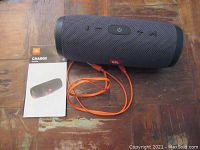 Photo of JBL Charge Essential portable Bluetooth speaker shown on wooden surface with an orange USB charging cable and user manual beside it.