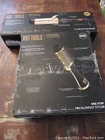 Two boxed Hot Tools styling products on wooden floor: 2-inch curling iron/wand and one-step pro blowout styler. Boxes show some wear and scuff marks.