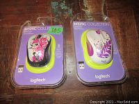 Front view of two Logitech M325C Collection wireless mice in original retail packaging on wooden surface. One mouse has pink and black floral design, the other purple floral design.
