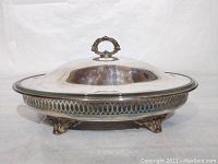 Side view of the silver plated covered serving dish closed, showing ornate handle and detailed edge with claw feet base.