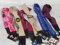 Six new guitar straps laid out on a white surface showing a variety of colors and materials (pink camo, tan with patch, beige, maroon, blue with pattern, solid maroon). All have black leather ends.