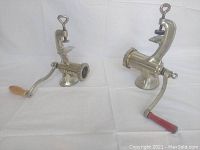 Two manual vintage meat grinders side-by-side on white surface; left grinder has wooden handle, right one has red handle and metal body.