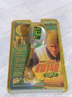 Front view of unopened Mini Real Swing Golf handheld game package showing game shaped like a golf club with LCD screen and keychain attached. Package features artwork of a male golfer and game features on front.