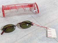 Side view of oval ELLE sunglasses with green lenses and metal frame, showing attached ELLE tag and clear case with red edging.