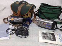 Overview of the two vintage camcorders (Sony and JVC), their respective carrying cases, power cords, and manuals laid out on a white surface.