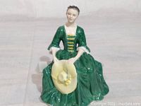 Front view of a seated female figurine in a green dress holding a yellow hat.