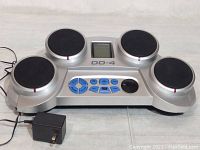 Front view of the Roland DD-4 electronic drum pad showing four black drum pads, control buttons, LCD screen, and power adapter.