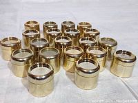 Front angled view of the set of 20 mini brass containers arranged on a white cloth background showing their shiny metallic brass finish and screw-on lids.