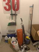 Photo showing Royal vintage upright vacuum with orange body, broom, bucket with cleaning supplies, and cleaning chemicals on floor.