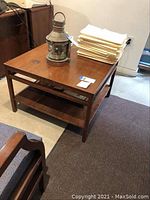 Front view of the square wood coffee table with reddish-brown finish, heavy and solid, showing an antique lantern and folded cloths on top.