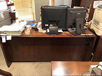 Full frontal view of executive desk showing top surface clutter with monitors and papers, back panel visible.