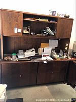Photo of the credenza and hutch showing the office furniture with dark brown wood finish and visible storage spaces, with papers and various small office items on the credenza surface.