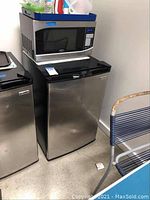 Mini fridge shows stainless steel door with black top and sides, a black handle, and a microwave placed on top of it.