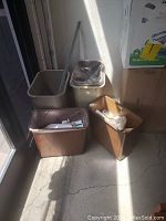 Five trash cans including beige plastic bins, brown rectangular bins, two with liners and some trash, placed near a door.