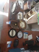 Various wall and desk clocks including a regulator pendulum wall clock with key, octagonal desk clock, a round black framed wall clock with large numbers, small round mirror, and framed duck print art on a wooden table.