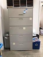 Full view of the metal vertical filing cabinet showing five drawers and the cabinet propped at an angle with a wooden strip under the base for stability.