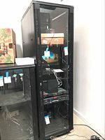 Tall metal server storage cabinet with glass front showing some cables and computer monitor inside. Black color and ventilation slots at sides of door.