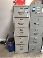 Front view of the gray metal filing cabinet showing the five drawers with handles and label holders, locking key visible on top drawer.