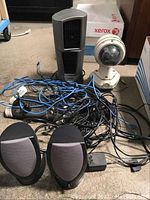 Items include a black and gray tower fan, white dome security camera, two black and gray speakers, and multiple assorted blue and black cords on a floor surface.