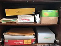 Boxes of clasp envelopes, gummed envelopes, security envelopes, and stacks of file pockets and paper on shelves in a wooden credenza.